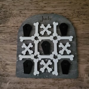 May include: A grey wooden game board with the name "Jude" engraved on it. The board features a tic-tac-toe style game with white bone-shaped pieces and black coffin-shaped pieces.