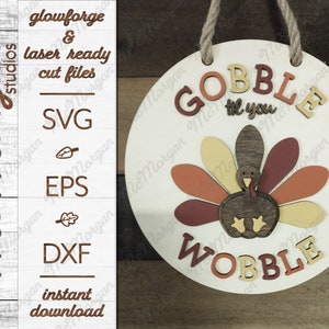 May include: A round wooden sign with the text "Gobble til you Wobble" in brown and yellow letters. A brown turkey with a red, orange, and yellow tail feathers is in the center of the sign.