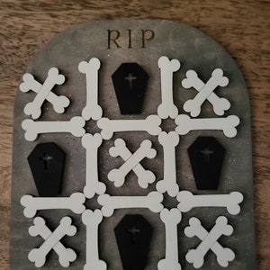 May include: A wooden game board with the word "RIP" at the top. The board is decorated with white bone shapes and black coffin shapes with crosses on them.