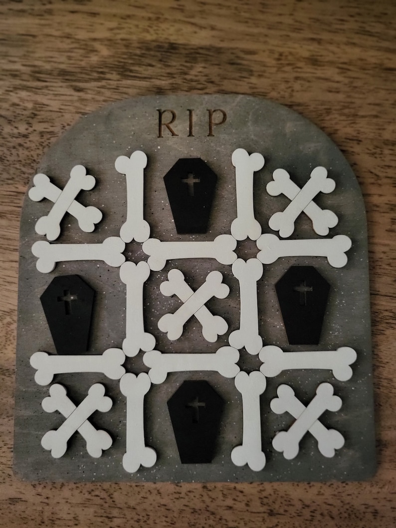 May include: A wooden game board with the word "RIP" at the top. The board is decorated with white bone shapes and black coffin shapes with crosses on them.