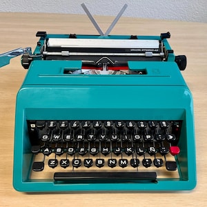 May include: A teal Olivetti Studio 46 typewriter with black keys and a red return key. The keys are arranged in a QWERTY layout. The paper holder is up. The typewriter sits on a light wood surface.