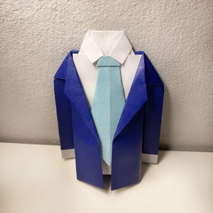 May include: Origami suit in blue, white, and light blue. The paper suit features a white collared shirt, a light blue tie, and a blue blazer. The origami is displayed on a white surface against a textured white background.
