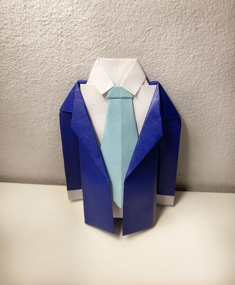 May include: Origami suit in blue, white, and light blue. The paper suit features a white collared shirt, a light blue tie, and a blue blazer. The origami is displayed on a white surface against a textured white background.