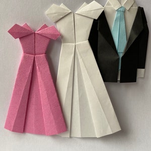 May include: Origami paper figures of a pink dress, a white dress, and a black suit with a light blue tie. The dresses have a pleated skirt design and off-the-shoulder neckline. The suit has a folded collar and lapels.