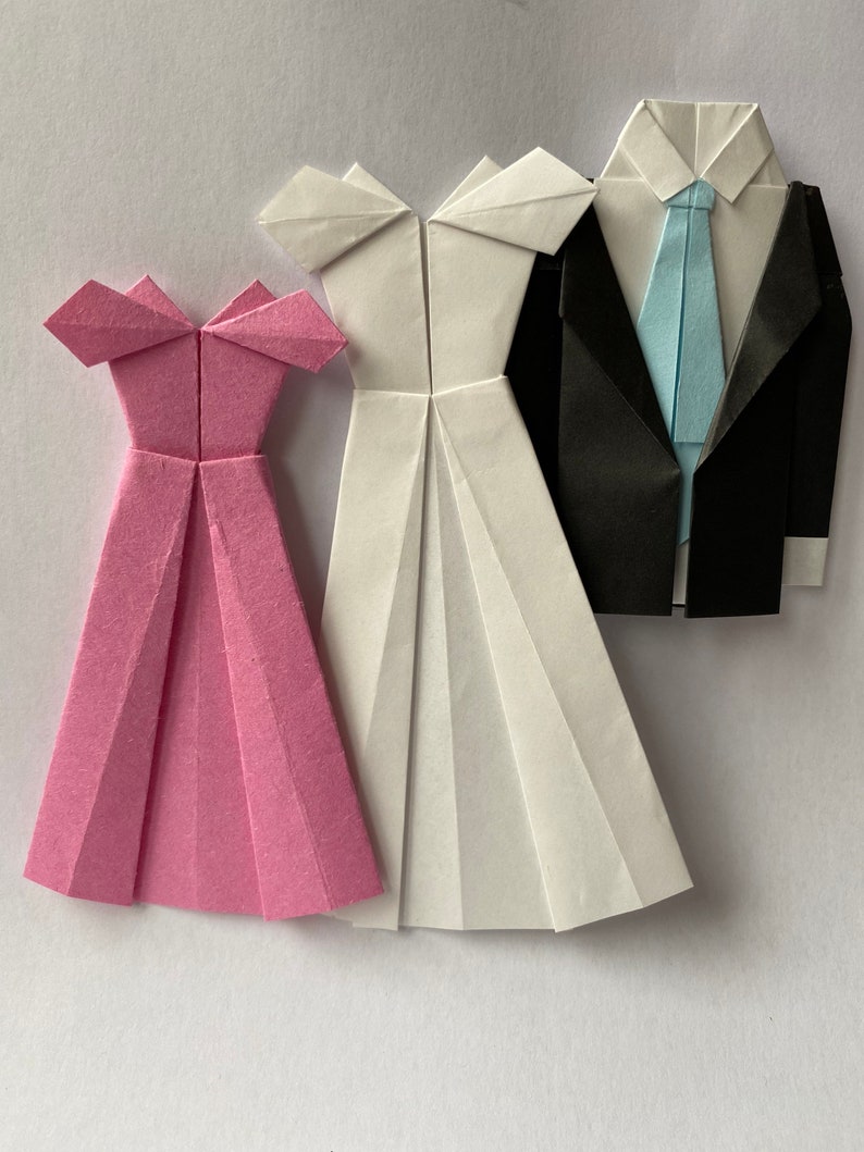 May include: Origami paper figures of a pink dress, a white dress, and a black suit with a light blue tie. The dresses have a pleated skirt design and off-the-shoulder neckline. The suit has a folded collar and lapels.