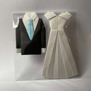May include: Origami wedding set featuring a black tuxedo with a light blue tie and a white wedding dress. The paper art is displayed in a clear plastic sleeve, perfect for a wedding or anniversary gift.