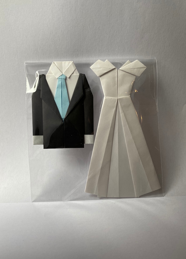 May include: Origami wedding set featuring a black tuxedo with a light blue tie and a white wedding dress. The paper art is displayed in a clear plastic sleeve, perfect for a wedding or anniversary gift.