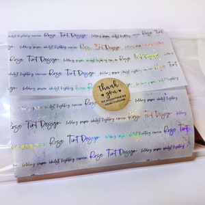May include: A clear plastic bag containing folded paper with holographic text and a small round sticker. The text on the paper reads "Rose Tint Design" and "folding paper whilst fighting cancer". The sticker says "thank you" and "for supporting my small business".