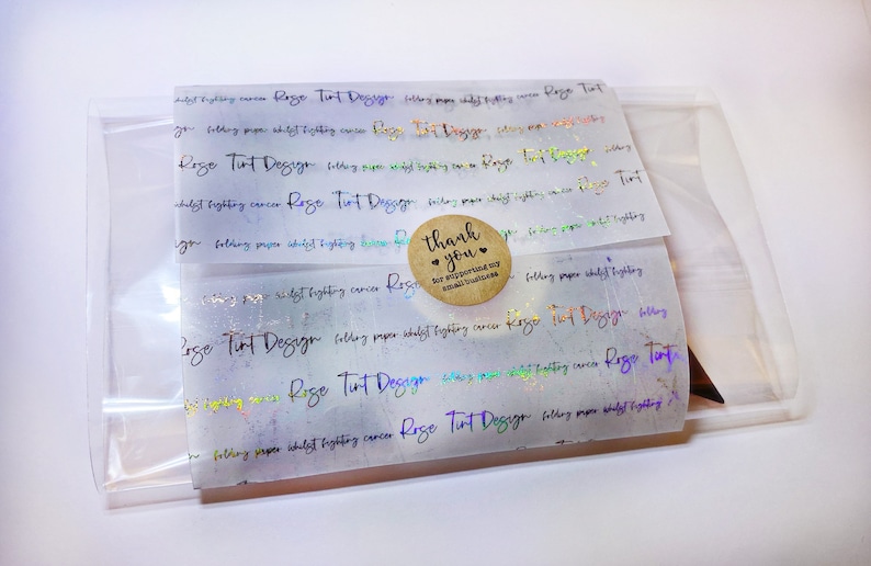 May include: A clear plastic bag containing folded paper with holographic text and a small round sticker. The text on the paper reads "Rose Tint Design" and "folding paper whilst fighting cancer". The sticker says "thank you" and "for supporting my small business".
