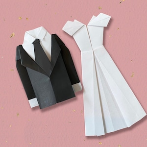 May include: Origami paper models of a tuxedo and a wedding dress. The black tuxedo features a white shirt and tie. The white dress has a pleated skirt and off-the-shoulder design. The background is pink with gold flecks.