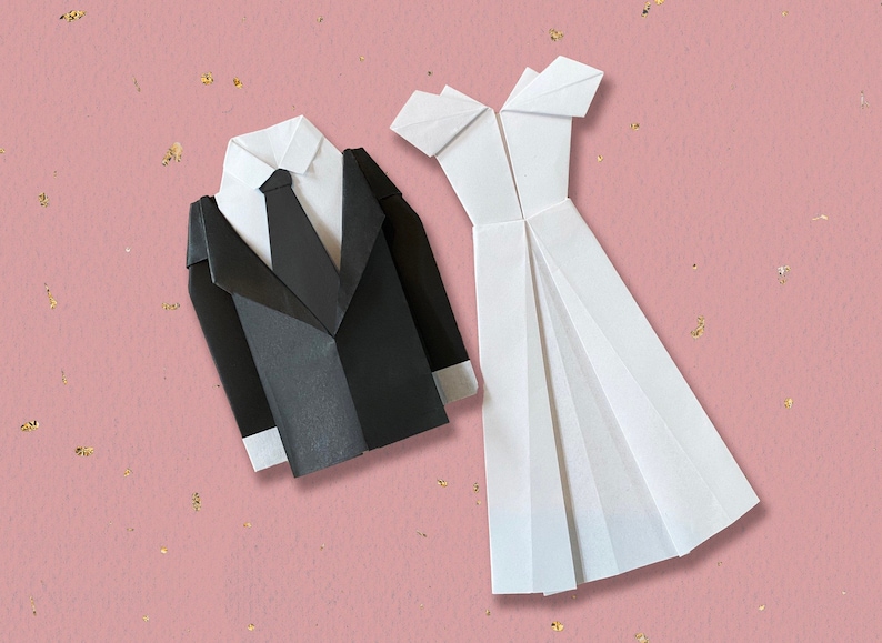 May include: Origami paper models of a tuxedo and a wedding dress. The black tuxedo features a white shirt and tie. The white dress has a pleated skirt and off-the-shoulder design. The background is pink with gold flecks.