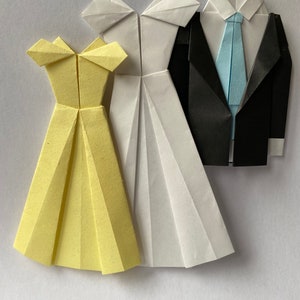 May include: Origami paper art featuring a yellow dress, a white dress, and a black suit with a light blue tie. The paper garments are folded with intricate detail, showcasing the art of paper folding. The dresses and suit are arranged on a white background.