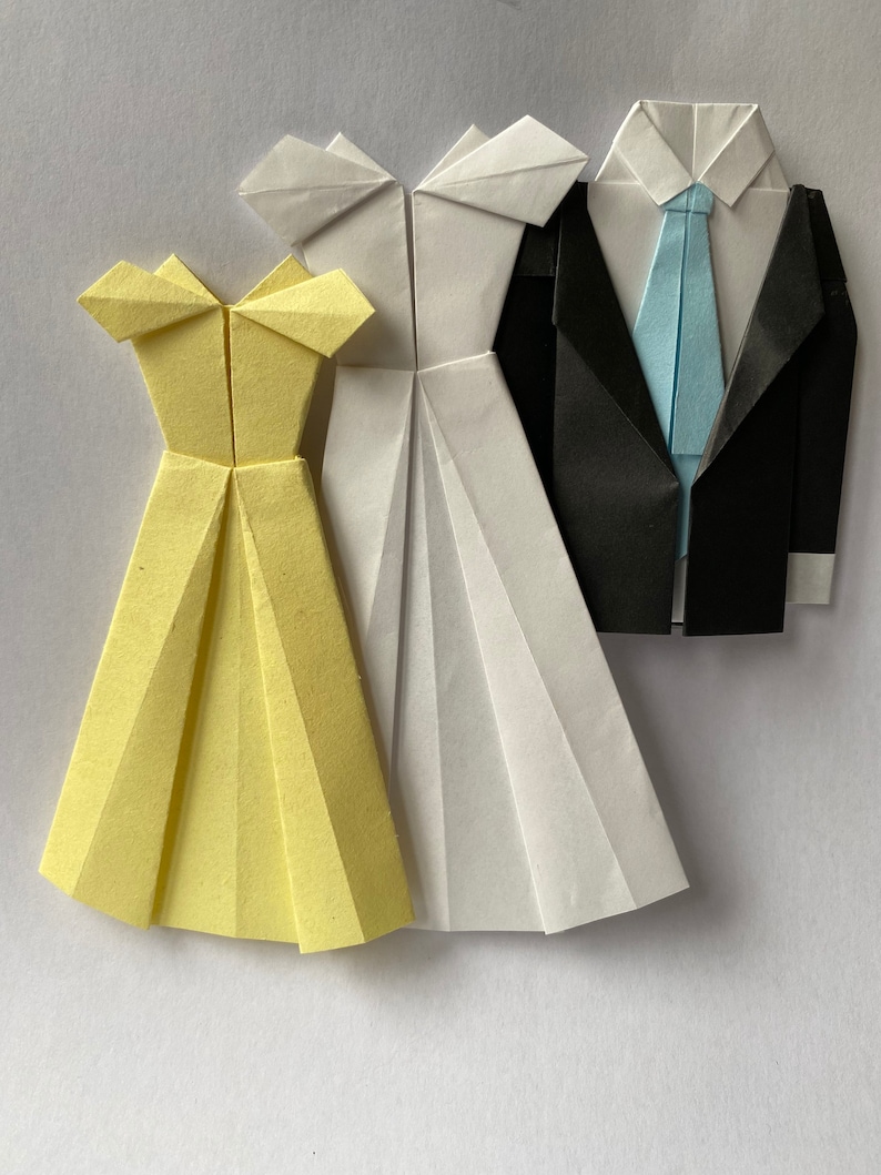 May include: Origami paper art featuring a yellow dress, a white dress, and a black suit with a light blue tie. The paper garments are folded with intricate detail, showcasing the art of paper folding. The dresses and suit are arranged on a white background.