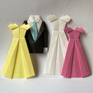 May include: Origami figures of a groom in a black suit with a blue tie, and three dresses. The dresses are yellow, white, and pink. The paper figures are arranged on a white background.