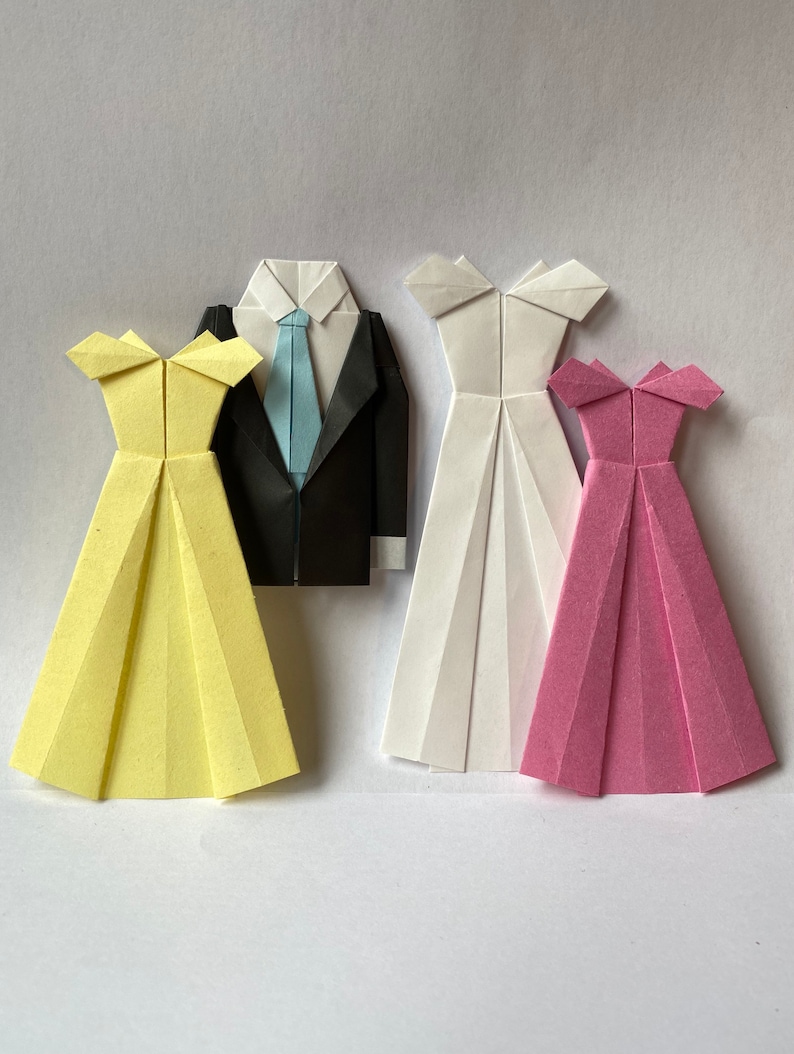 May include: Origami figures of a groom in a black suit with a blue tie, and three dresses. The dresses are yellow, white, and pink. The paper figures are arranged on a white background.