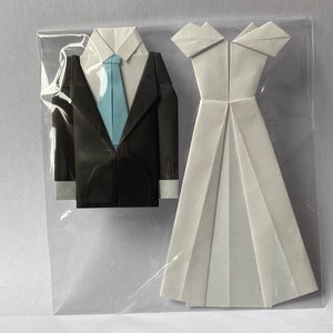 May include: Origami wedding set featuring a black tuxedo with a light blue tie and a white wedding dress. The paper art is displayed in a clear plastic sleeve, showcasing the folded paper details.