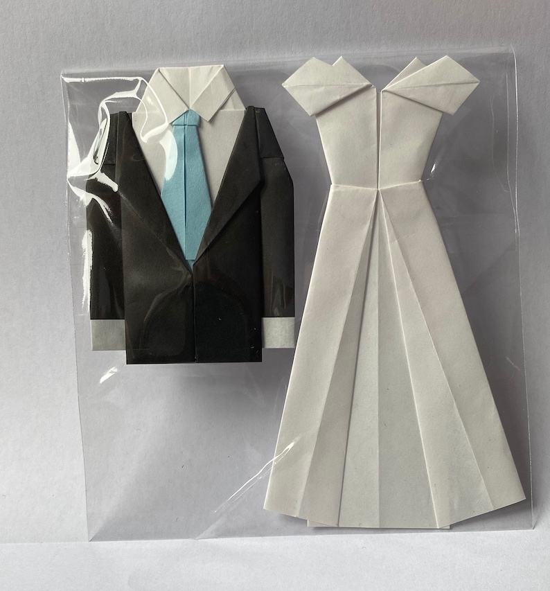 May include: Origami wedding set featuring a black tuxedo with a light blue tie and a white wedding dress. The paper art is displayed in a clear plastic sleeve, showcasing the folded paper details.
