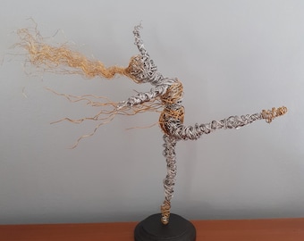 May include: A wire sculpture depicting a ballerina in a poised stance. The figure is made from silver and gold-toned wire, with flowing strands suggesting hair. The sculpture is mounted on a small, circular, black base, creating a dynamic and artistic display.