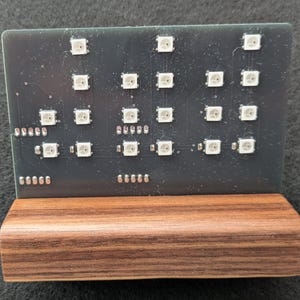 May include: A black circuit board with 16 white LED lights arranged in a grid pattern. The board is mounted on a wooden stand.
