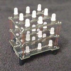 May include: A small, three-dimensional electronic component featuring multiple white LED lights arranged in a cube shape. The LEDs are mounted on circuit boards, connected by copper wiring, and supported by black feet.