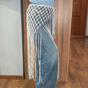 Könnte beinhalten: Ein cremefarbener Makramee-Gürtel mit langen Fransen. Der Gürtel wird über einer blauen Jeans getragen. Das Makramee-Design weist ein geometrisches Muster auf. Der Gürtel besteht aus einem weichen, gewebten Material.