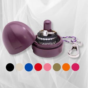 Puede incluir: Un estuche para anillos en forma de huevo de color ciruela con un clip metálico plateado extraíble y un cordón morado estampado. El estuche abierto muestra un poste central con tres anillos apilados, uno con piedra rectangular.