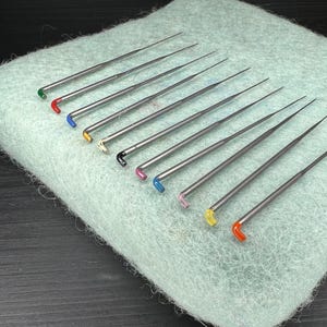 May include: A collection of needle felting tools with colorful handles, including green, red, blue, yellow, black, and pink. The needles are arranged on a light green felted wool surface, showcasing the variety of colors and tools.