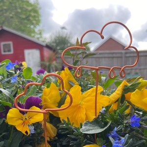May include: Two copper wire garden stakes shaped like clouds with raindrops hanging from the bottom. The stakes are placed in a flower pot with yellow and purple flowers.