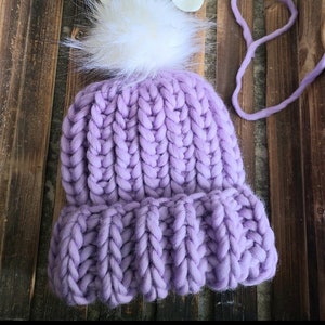 May include: A purple knitted beanie with a white pom pom. The beanie is made of chunky yarn and has a ribbed texture.
