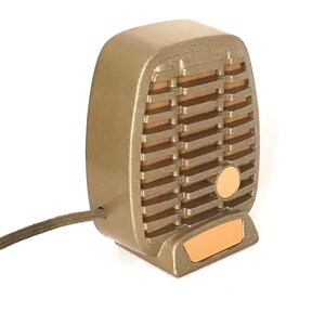 Puede incluir: Un micr&oacute;fono de estilo vintage en tonos dorados con un dise&ntilde;o de rejilla rectangular, lamas horizontales, un acento circular y una peque&ntilde;a placa base rectangular. Un cable fino recubierto de tela oscura sale del lateral del dispositivo.