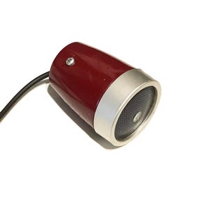 May include: A cylindrical speaker with a glossy dark red body and a silver metallic front rim. The front face features a black perforated grille with a small circular center detail. A black electrical cord extends from the rear of the unit.