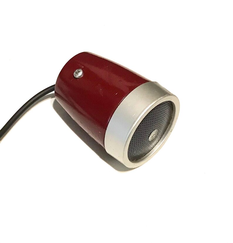 May include: A cylindrical speaker with a glossy dark red body and a silver metallic front rim. The front face features a black perforated grille with a small circular center detail. A black electrical cord extends from the rear of the unit.