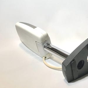 May include: A white rectangular microphone with a silver mesh grille, attached to a chrome-plated metal arm and a black textured mounting base. A white power cord extends from the base of the microphone housing, which features embossed text.