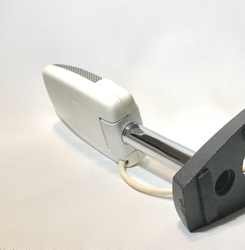 May include: A white rectangular microphone with a silver mesh grille, attached to a chrome-plated metal arm and a black textured mounting base. A white power cord extends from the base of the microphone housing, which features embossed text.