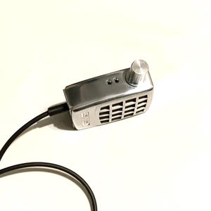 Puede incluir: Un dispositivo electr&oacute;nico rectangular de color plateado con acabado met&aacute;lico, que presenta una perilla de control circular en la parte superior derecha, una rejilla de altavoz, dos puertos peque&ntilde;os y un cable de alimentaci&oacute;n negro a la izquierda.