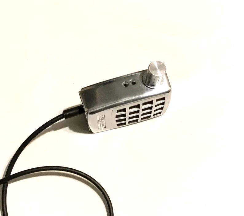Puede incluir: Un dispositivo electr&oacute;nico rectangular de color plateado con acabado met&aacute;lico, que presenta una perilla de control circular en la parte superior derecha, una rejilla de altavoz, dos puertos peque&ntilde;os y un cable de alimentaci&oacute;n negro a la izquierda.
