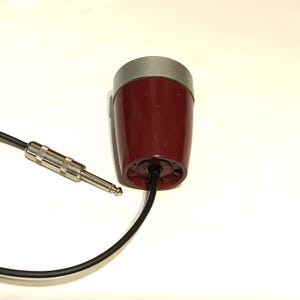 May include: A maroon, cylindrical microphone with a silver top cap and a black cable attached to the base. The cable features a standard silver-colored metal audio jack connector, which is used for connecting the device to audio equipment.
