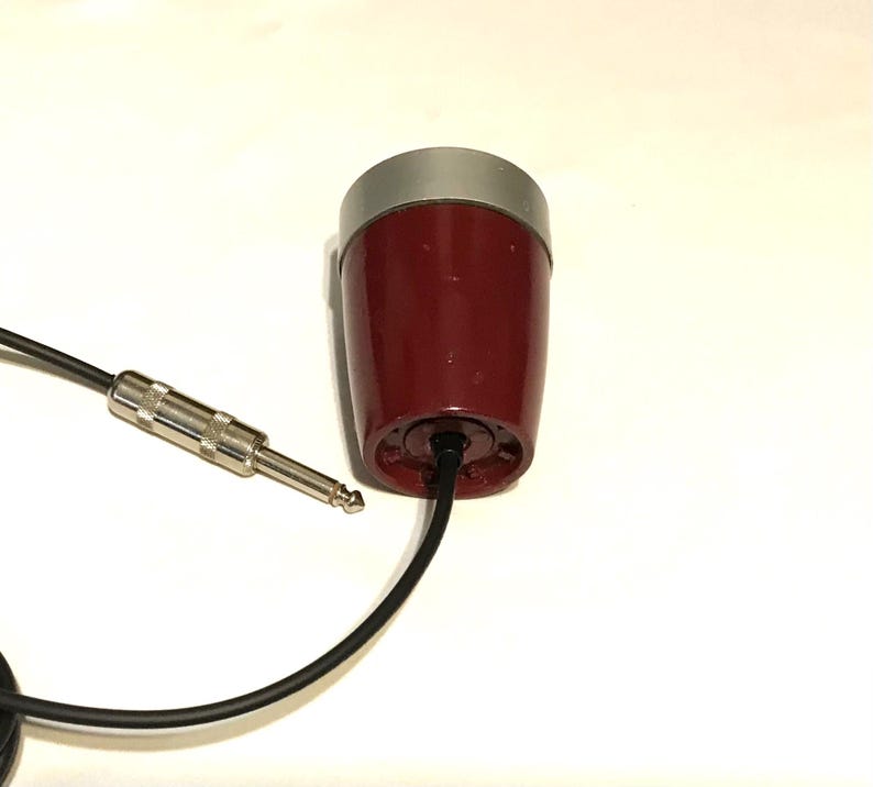 May include: A maroon, cylindrical microphone with a silver top cap and a black cable attached to the base. The cable features a standard silver-colored metal audio jack connector, which is used for connecting the device to audio equipment.