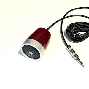 May include: A vintage-style microphone with a deep red, tapered cylindrical body and a silver-toned circular front grille. The microphone is attached to a long black cable that terminates in a standard silver-colored quarter-inch audio jack plug.