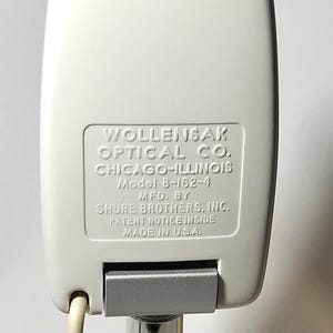 May include: A white microphone with a rectangular housing featuring embossed text: WOLLENSAK OPTICAL CO. CHICAGO-ILLINOIS Model B-162-4 MFD. BY SHURE BROTHERS, INC. PATENT NOTICE INSIDE MADE IN U.S.A. A silver metallic hinge is visible at the base.