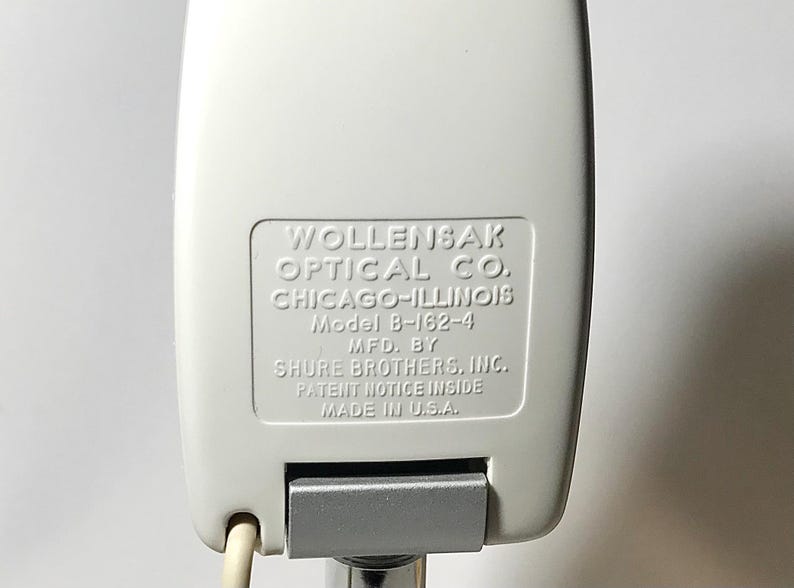 May include: A white microphone with a rectangular housing featuring embossed text: WOLLENSAK OPTICAL CO. CHICAGO-ILLINOIS Model B-162-4 MFD. BY SHURE BROTHERS, INC. PATENT NOTICE INSIDE MADE IN U.S.A. A silver metallic hinge is visible at the base.