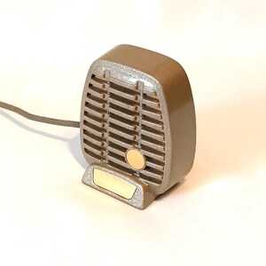 Puede incluir: Un micr&oacute;fono de estilo vintage en color topo met&aacute;lico con una rejilla rectangular, un detalle circular en tono dorado y una placa base rectangular dorada. Un cable gris fino se extiende desde el lado izquierdo del cuerpo del micr&oacute;fono.