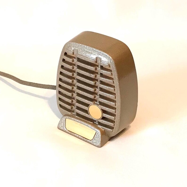 Puede incluir: Un micr&oacute;fono de estilo vintage en color topo met&aacute;lico con una rejilla rectangular, un detalle circular en tono dorado y una placa base rectangular dorada. Un cable gris fino se extiende desde el lado izquierdo del cuerpo del micr&oacute;fono.
