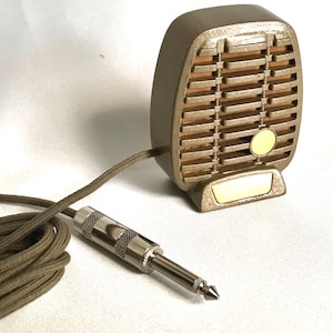 Puede incluir: Un micr&oacute;fono de estilo vintage en tono dorado con rejilla texturizada, fondo de tela beige y un peque&ntilde;o emblema circular. Cuenta con una base dorada a juego y est&aacute; conectado a un cable largo trenzado en color beige con un conector jack plateado.