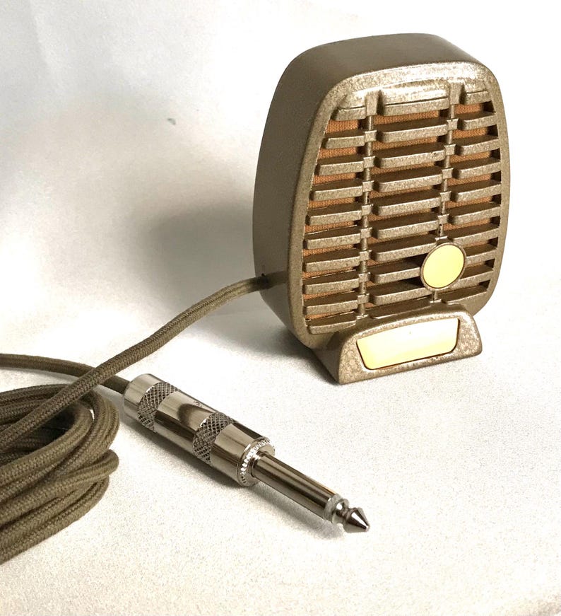 Puede incluir: Un micr&oacute;fono de estilo vintage en tono dorado con rejilla texturizada, fondo de tela beige y un peque&ntilde;o emblema circular. Cuenta con una base dorada a juego y est&aacute; conectado a un cable largo trenzado en color beige con un conector jack plateado.