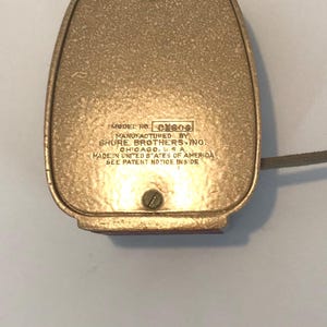 Puede incluir: Una base de micr&oacute;fono de metal dorado con acabado texturizado. El texto dice: MODEL NO. CX80G, MANUFACTURED BY SHURE BROTHERS INC. CHICAGO, U.S.A., MADE IN UNITED STATES OF AMERICA, SEE PATENT NOTICE INSIDE. Se observa un tornillo.