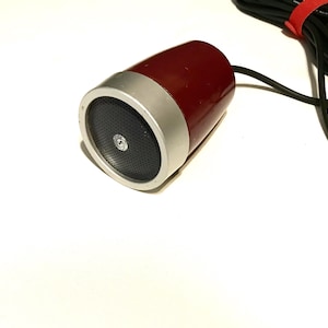 May include: A cylindrical microphone with a deep red body and a silver metallic front grille. The front features a black perforated speaker mesh with a small circular logo in the center. A black cable is attached to the rear with a red velcro tie.