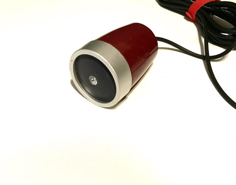May include: A cylindrical microphone with a deep red body and a silver metallic front grille. The front features a black perforated speaker mesh with a small circular logo in the center. A black cable is attached to the rear with a red velcro tie.