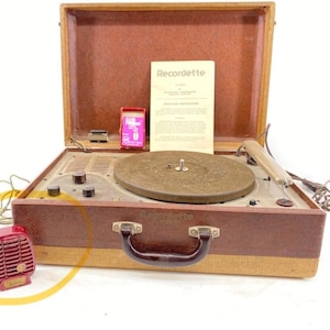 Puede incluir: Un tocadiscos port&aacute;til vintage Recordette en un malet&iacute;n marr&oacute;n y beige. Incluye un plato giratorio circular, perillas de control, un brazo fonocaptor, un micr&oacute;fono rojo, una peque&ntilde;a caja rosa y un manual de instrucciones marcado Recordette.