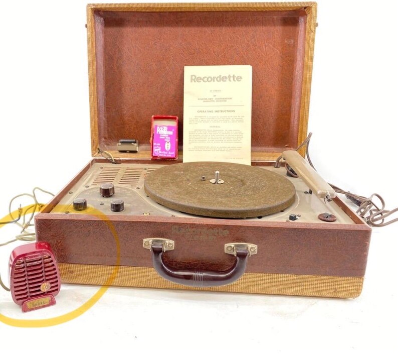 Puede incluir: Un tocadiscos port&aacute;til vintage Recordette en un malet&iacute;n marr&oacute;n y beige. Incluye un plato giratorio circular, perillas de control, un brazo fonocaptor, un micr&oacute;fono rojo, una peque&ntilde;a caja rosa y un manual de instrucciones marcado Recordette.